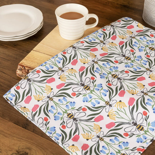 A floral tea towel with a design of bees and flowers, laid out on a wooden surface with a cup of tea and plates in the background.